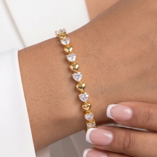 A close-up of a gold heart-link bracelet with clear heart-shaped stones worn on a woman's wrist, styled with a white blazer and manicured nails.